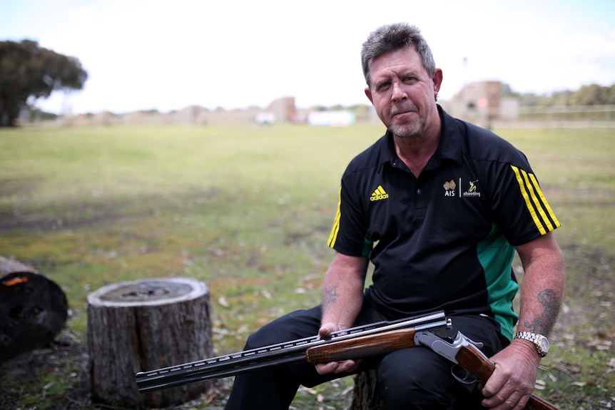 A man in a polo shirt sits on a stump with a cocked gun.