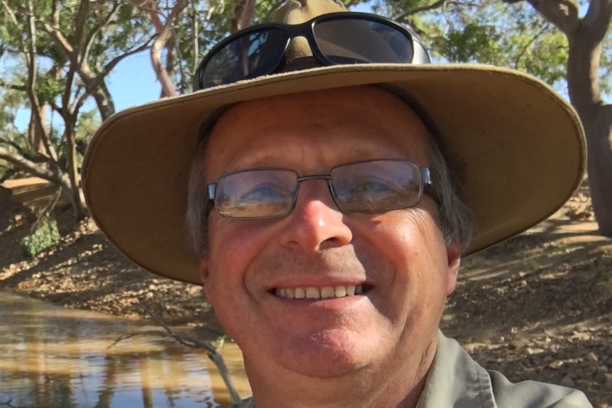 A smiling middle-aged man wearing square-framed glasses and a brown hat and sunglasses by a river bed. 
