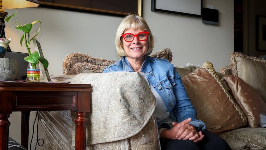 A woman wearing bright red glasses sits on a couch next to a wooden side table and house plant.