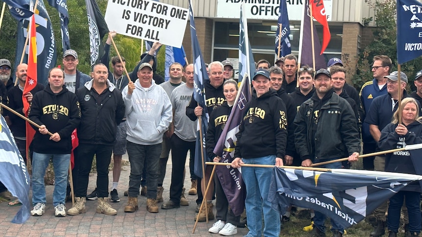 A group of workers in union-branded clothing stand holding flags in front of an office at an industrial site.