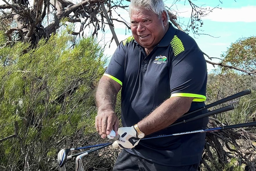 A man smiling holding a ball and golf sticks in from of trees