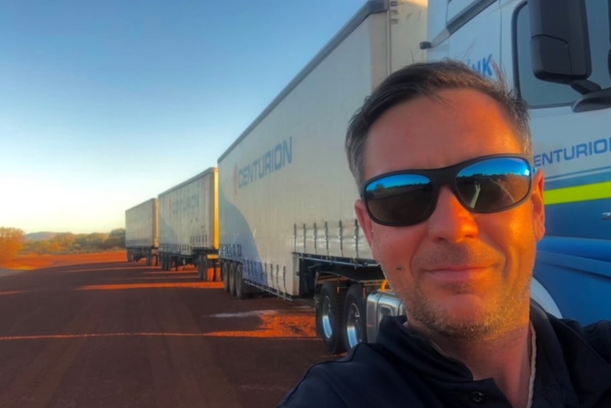 A man with glasses on on a dirt road with a truck in the background