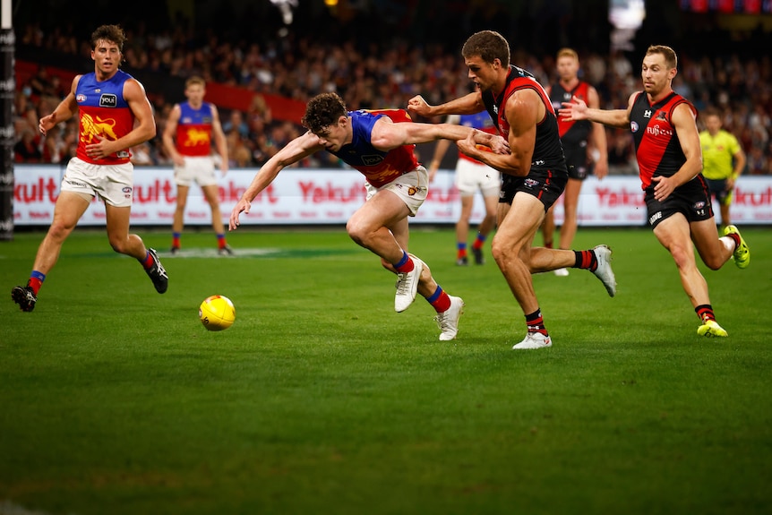 An AFL player puts his head down and reaches for the ball ahead of him, while a defender tries to hold his arm back.
