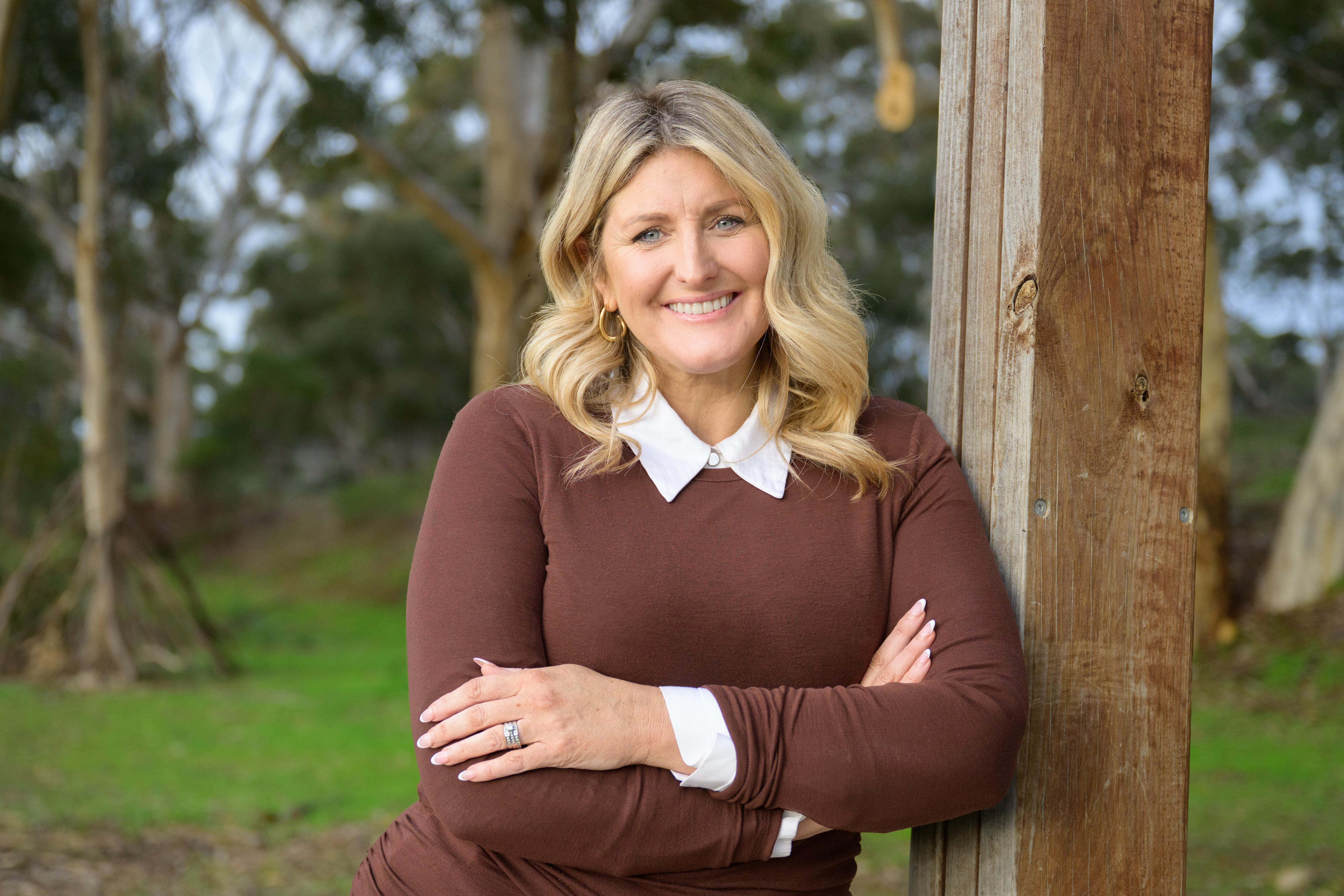 A smiling blonde woman in a brown jumper leans against a wooden post.