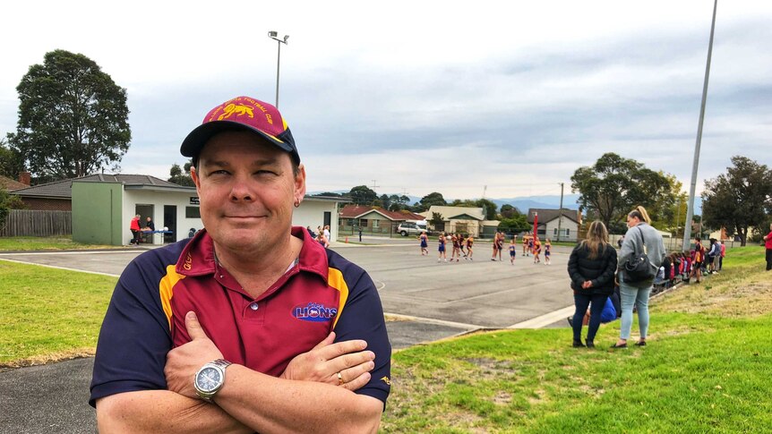Moe Junior Football Club president Mark Walsh stands with his arms folded with a netball game being played in the background.