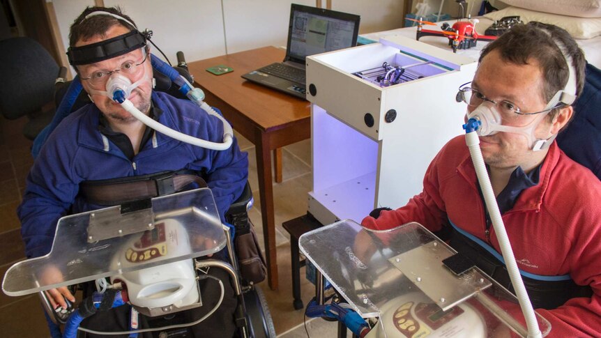 Two men in wheelchairs with breathing apparatus sit next to a 3D printer