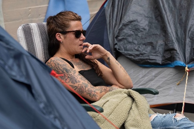 A woman wearing sunglasses sits on a camp chair outside a tent and looks away.