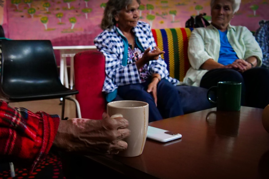 An old woman holds a coffee cup at a community table.