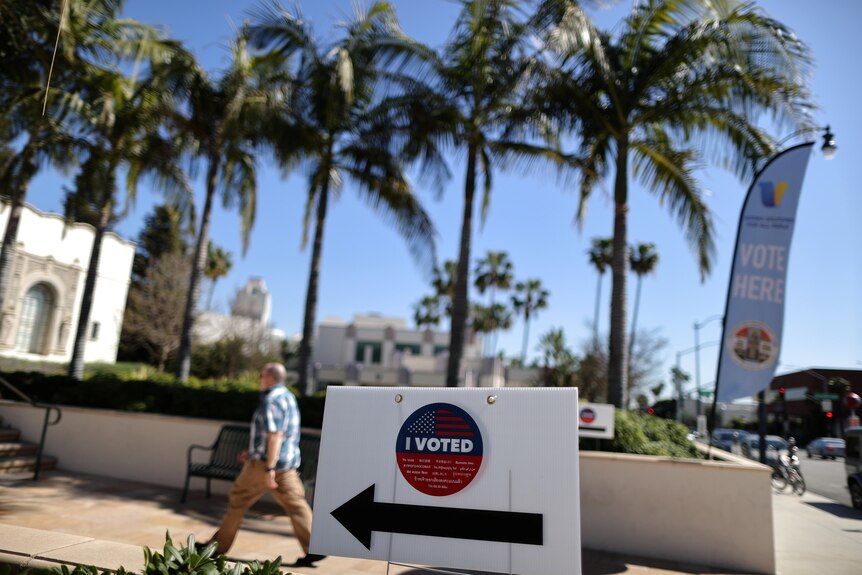 A middle-aged man walks past a row of palm trees in the direction pointed to by a sign saying "I voted".