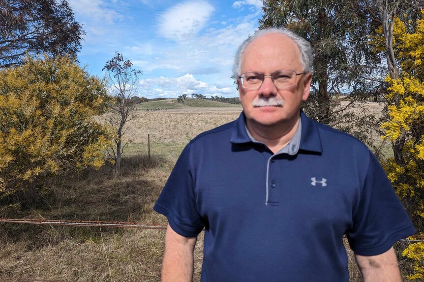 Man with glasses and moustache wearing a blue shirt standing beside paddock of dry grass. 