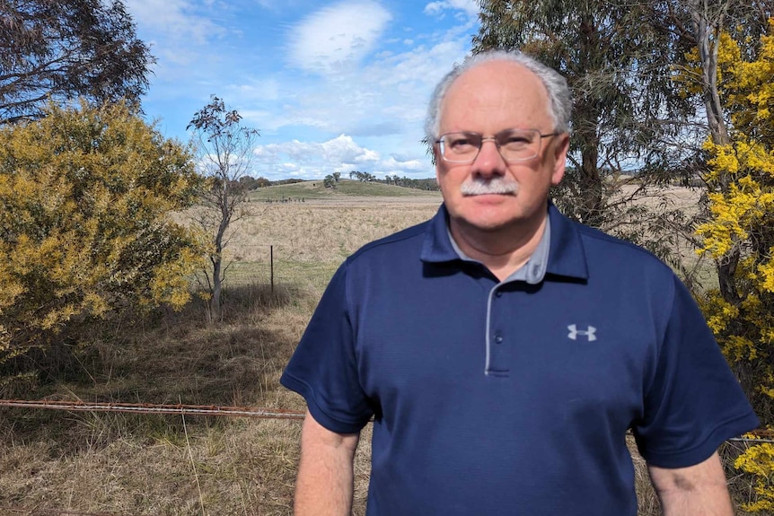 Man with glasses and moustache wearing a blue shirt standing beside paddock of dry grass. 