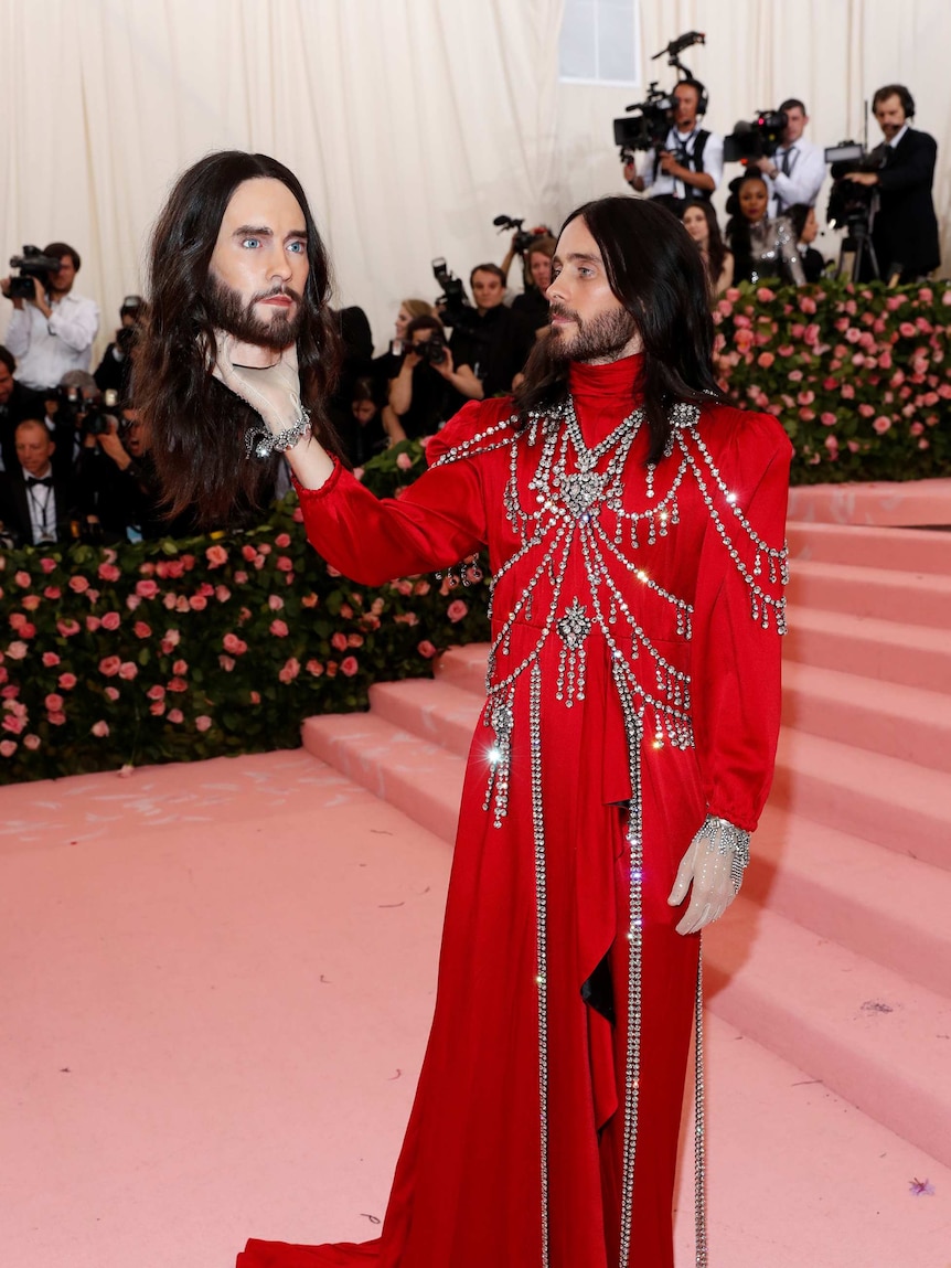 Jared Leto in a red gown, holing a mould of his own head