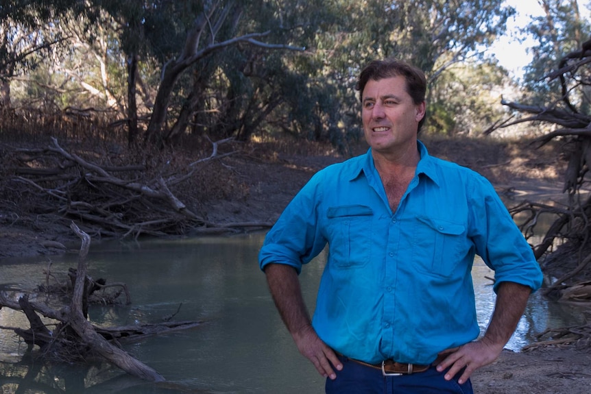 a man stands with his hands on his hips, before a river that looks low on water, surrounded by sticks