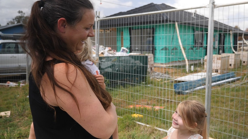 A woman stands outside a home being constructed, with two small children.