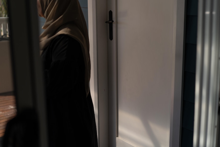 A woman in Islamic garments is shown from the side, standing in a doorway, looking outdoors.