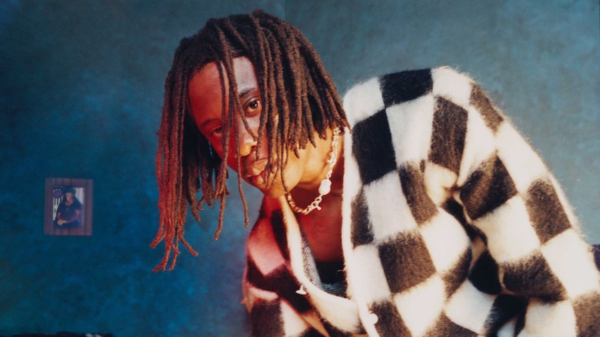 Obongjayar has dreadlocked hair and wears a black and white checkerboard pattern sweater. The background is blue.