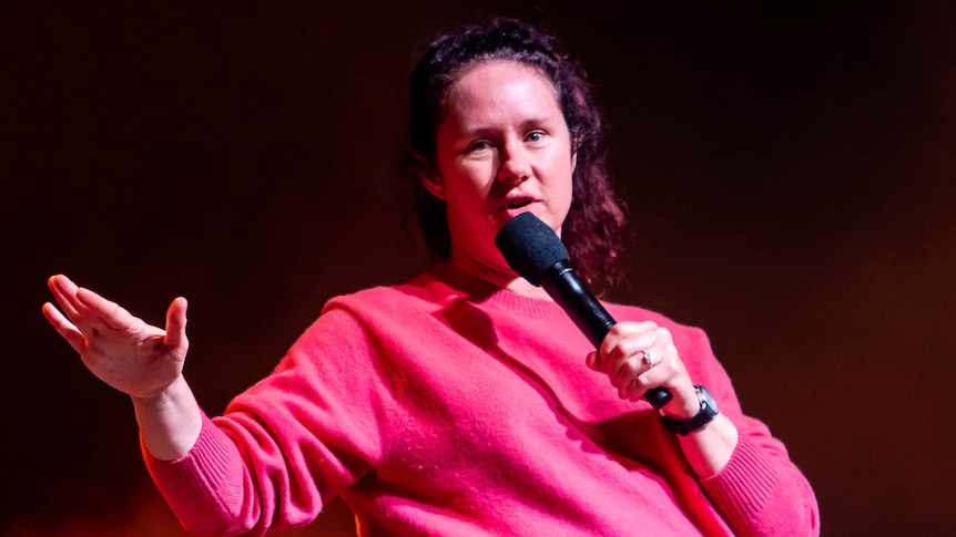 Mel Buttle, a 30-something woman, with her hair in a high pony and wearing a pink sweater, speaks into a microphone on stage 