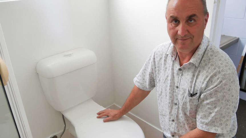 Chris kneels next to a toilet seat, and looks up at the camera.