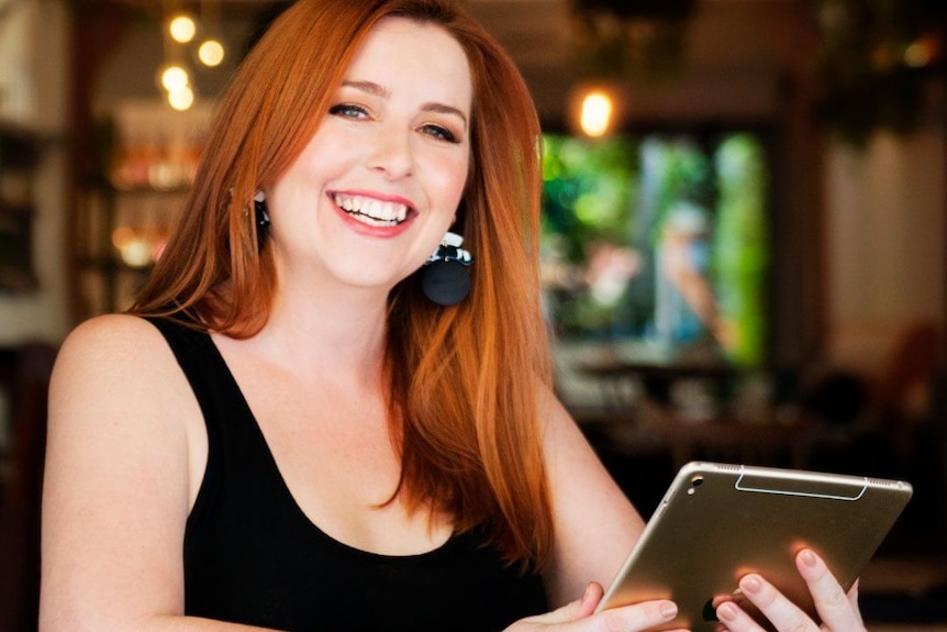 A woman with red hair smiles holding an iPad.
