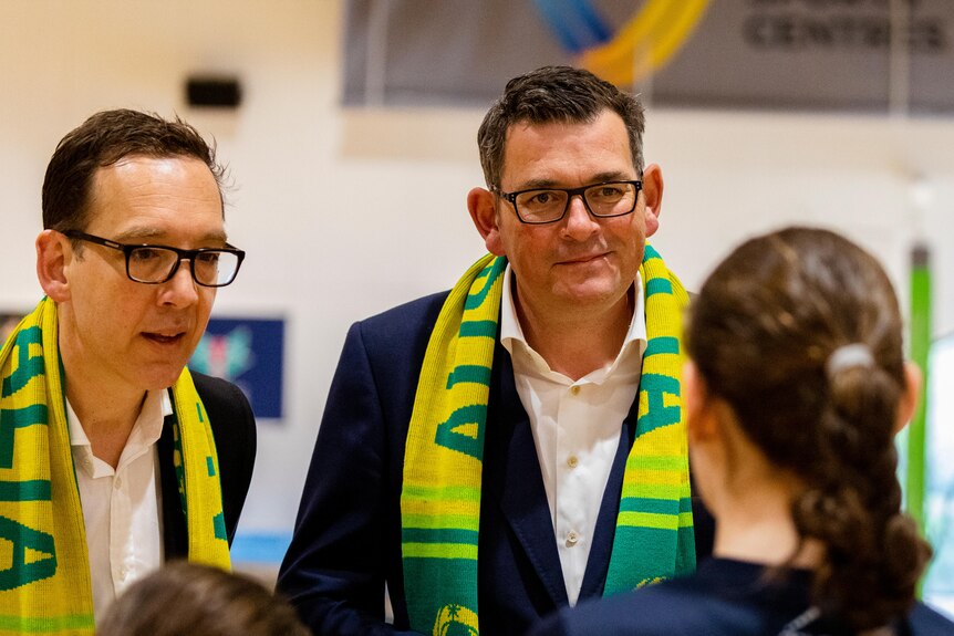 The Premier and Steve Dimopoulos smile, dressed in green and gold in a netball stadium.