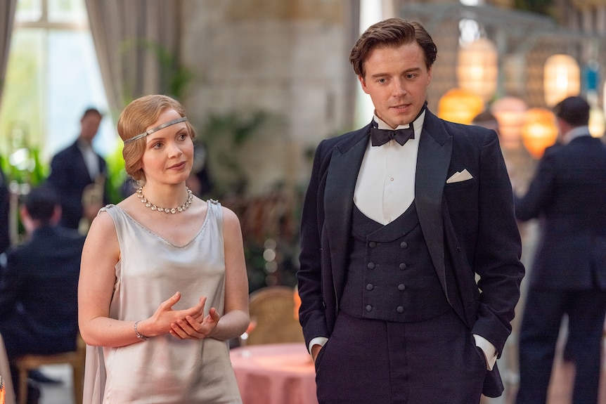 White woman with blonde 1940s-styled hair wears white silk dress and looks at white man with brown hair in black tuxedo.