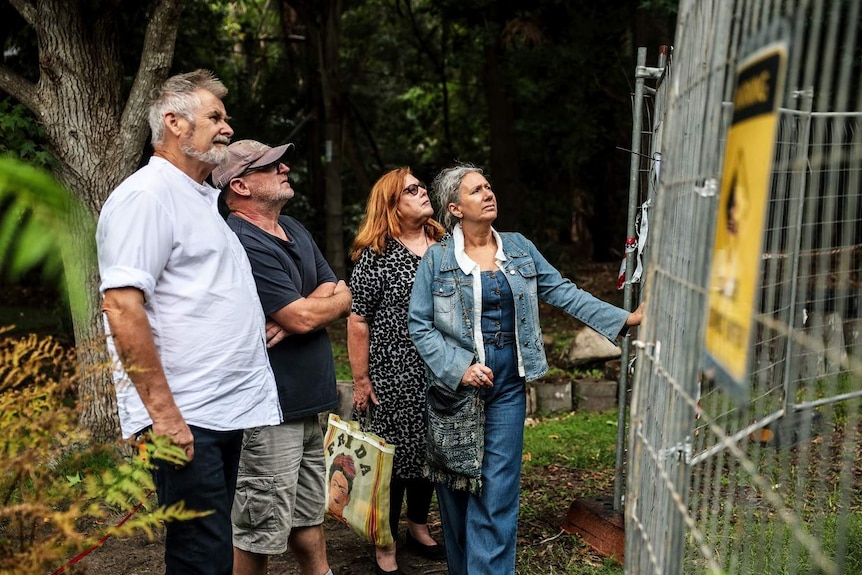 Two men and two women stand next to a temporary fence looking unhappy. A yellow danger sign is out of focus in the foreground.