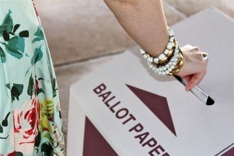 An unidentified woman puts her vote into ballot box, arm has bracelets, light green dress with orange, pink roses can be seen.