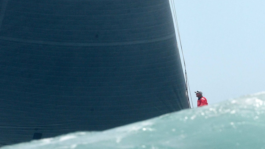 Image composed of a wave, the mainsail and a crew member of Wild Oats XI - photographed over the top of a wave.