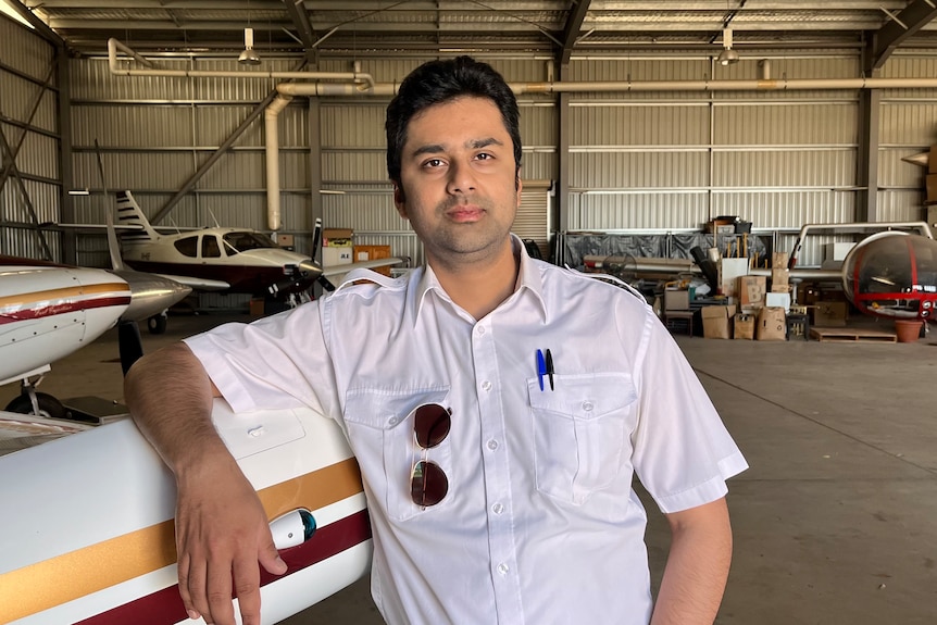 A serious young Indian man wears white shirt, sun glasses in pocket, pen in another, leans on an small airplane in a hangar.