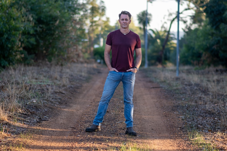 Jarrod wears a red tshirt and blue jeans standing on a dirt driveway among bushland and tall trees.