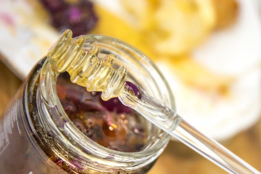 A close-up of a honey stick resting on the top of a glass jar of honey with dried rose petals in it.