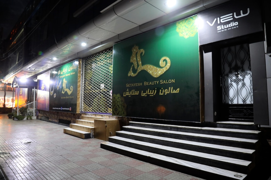 a row of closed shops in the night with green signage saying beauty salon and metal fence in front of the door 