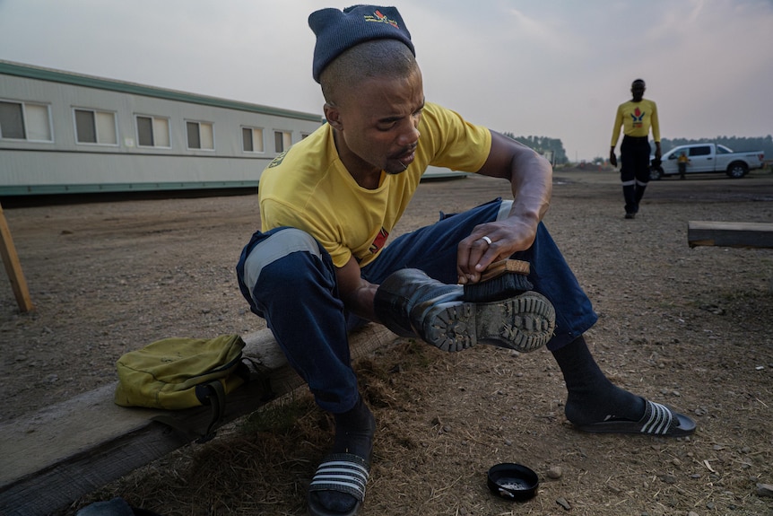 A South African firefighter in a yellow t-shirt polishes his fire boots while sitting on a low bench.