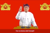 An elderly South-East Asian with glasses in traditional white tunic and red and blue hat raises three fingers at lecturn.