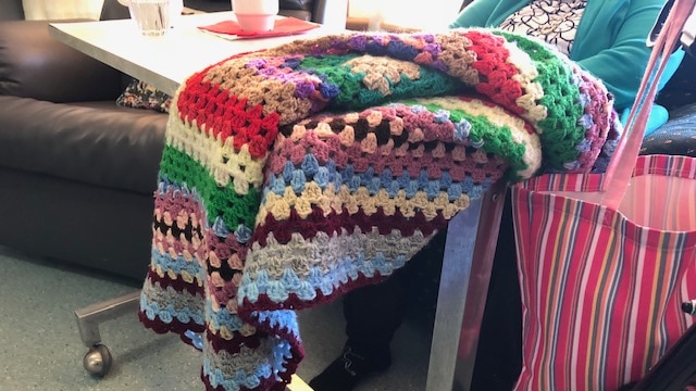 A crocheted blanket hangs over a food trolley pushed up to a resident (obscured) in a chair at a nursing home.