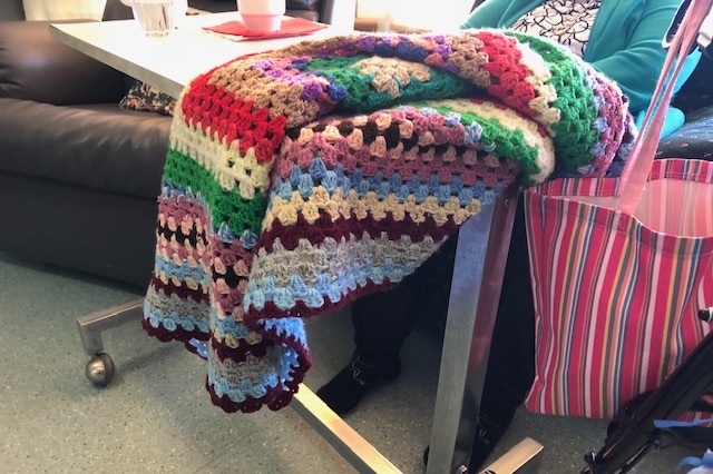 A crocheted blanket hangs over a food trolley pushed up to a resident (obscured) in a chair at a nursing home.