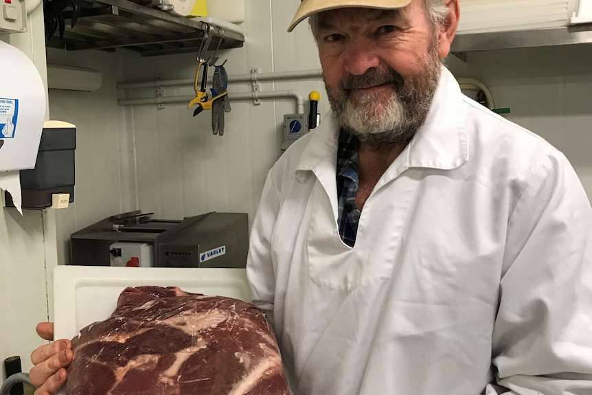 A grey-bearded man in a green cap and white butcher's coat holds a tray of steaks aloft