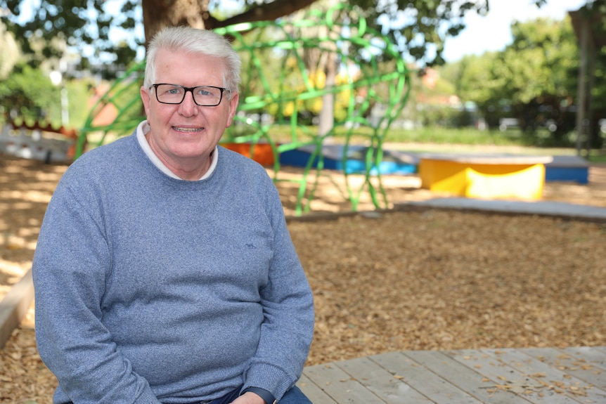 A man wearing glasses and a blue jumper sits with a playground behind him.
