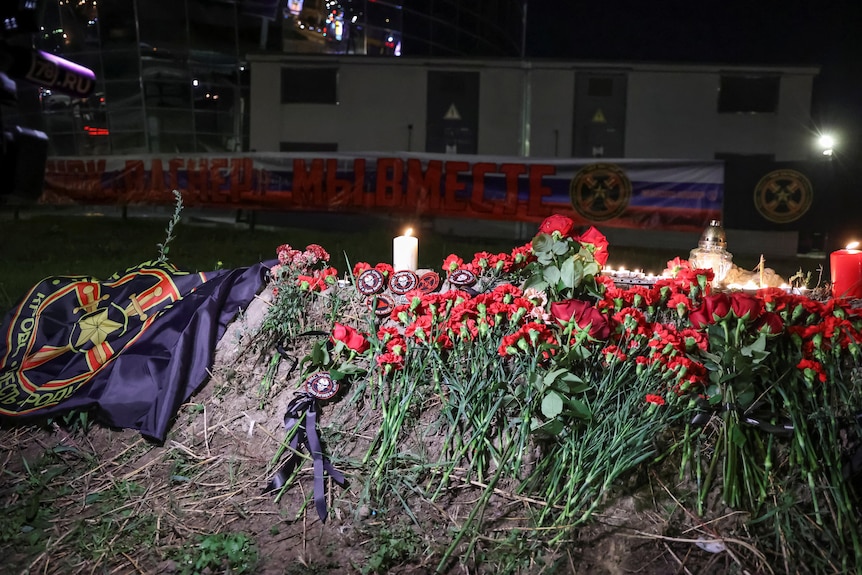 A pile of flowers, with candles and a flag, at night.