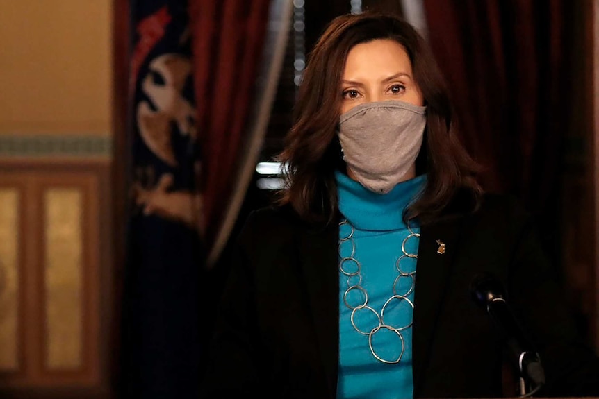 Gretchen Whitmer wearing a mask speaks to camera with a flag behind her