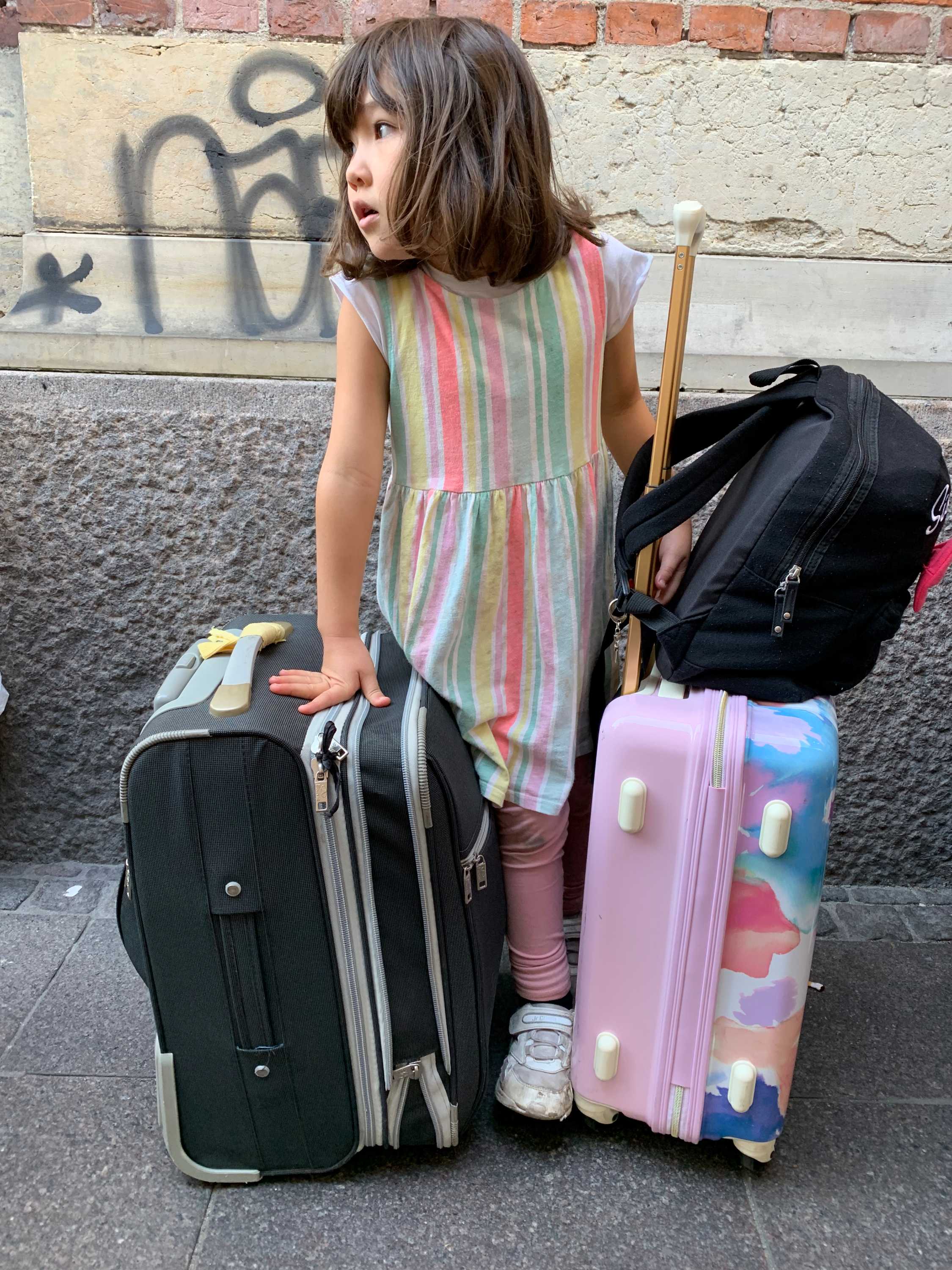 A young girl stand in front of a brick wall with several pieces of luggage.