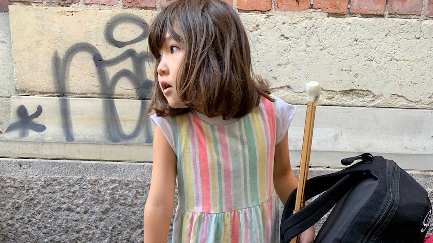 A young girl stand in front of a brick wall with several pieces of luggage.