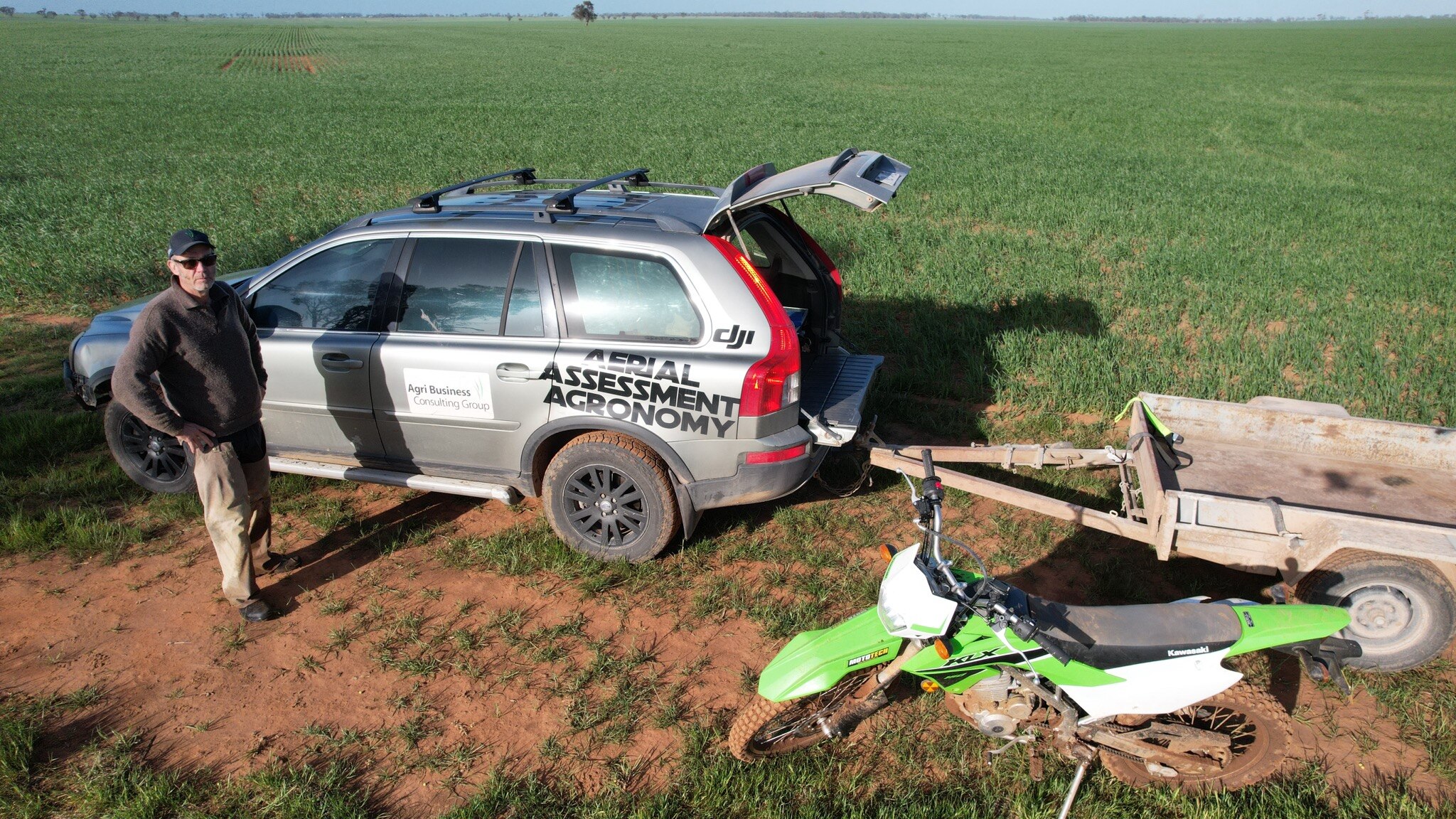 A farmer with sunglasses on looking up at a drone image standing in a field in front of a car, trailer and motorbike.