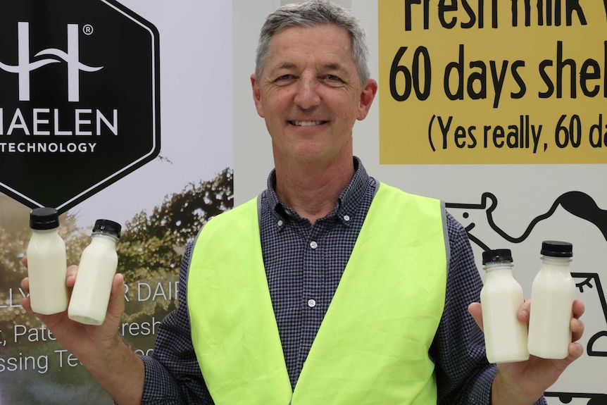 A man in a fluoro vest holds up four small bottles of unlabelled milk in front of a sign saying Halen Technology.