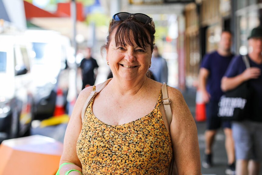 A lady with sunglasses and a floral top smiles on the street 