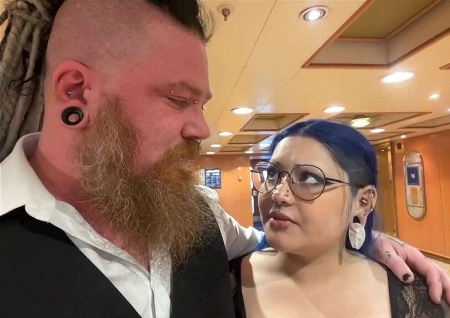 A man with an earring, beard, braids and a shaved side of the head looks at a woman with purple hair and earrings