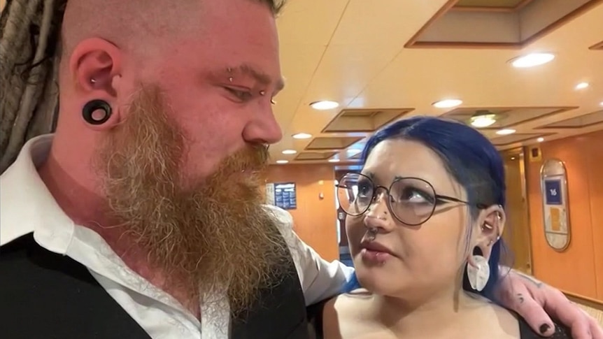 A man with an earring, beard, braids and a shaved side of the head looks at a woman with purple hair and earrings