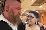 A man with an earring, beard, braids and a shaved side of the head looks at a woman with purple hair and earrings