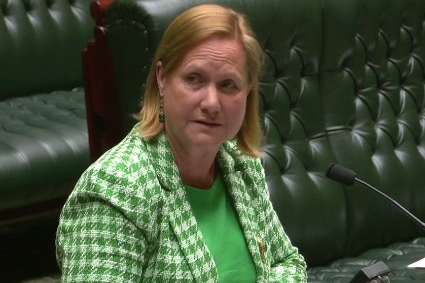 Woman in green suit with blonde hair speaks into microphone with green chairs behind her.