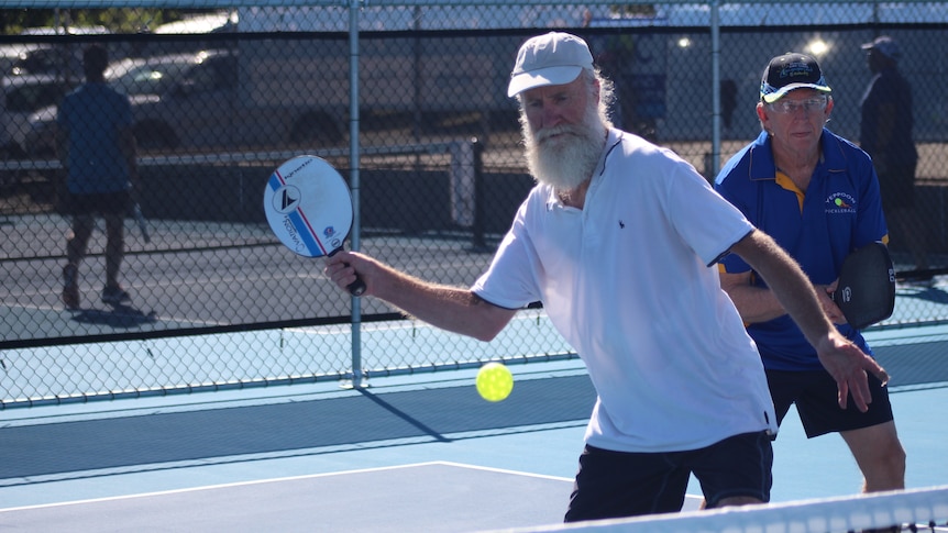Two older men playing pickleball with one man attempting to strike the plastic ball with his paddle.